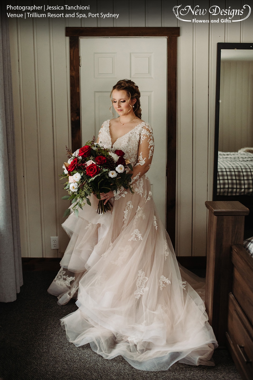 A bride stands in a Chalet Room, her dress swirls around her, and she holds a bouquet of lush winter greens, Bright red and white flowers, with accenting burgundy miniature roses, berries, and pinecones